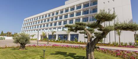 A modern hotel with a symmetrical facade, multiple balconies, and a well-maintained garden with a prominent tree.