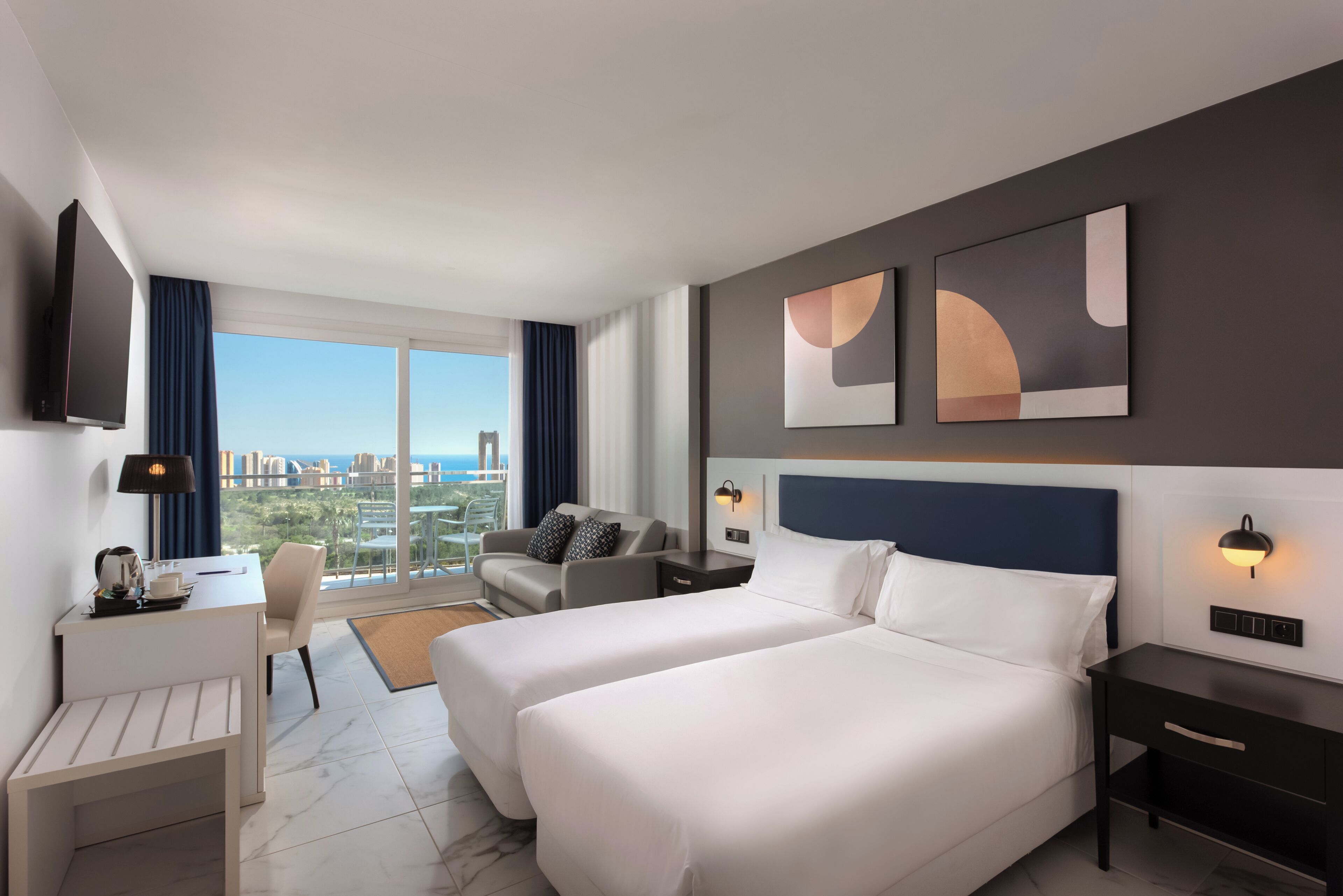 Ein Hotelzimmer mit zwei Betten, einem Schreibtisch, einem Sessel, einem Fernseher und Blick auf die Stadt.