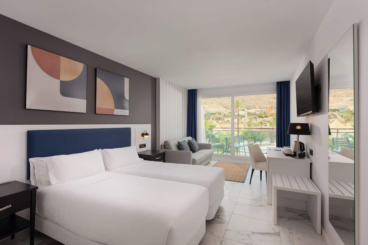 Habitación de hotel con cama, escritorio, silla, televisor y balcón con vistas.