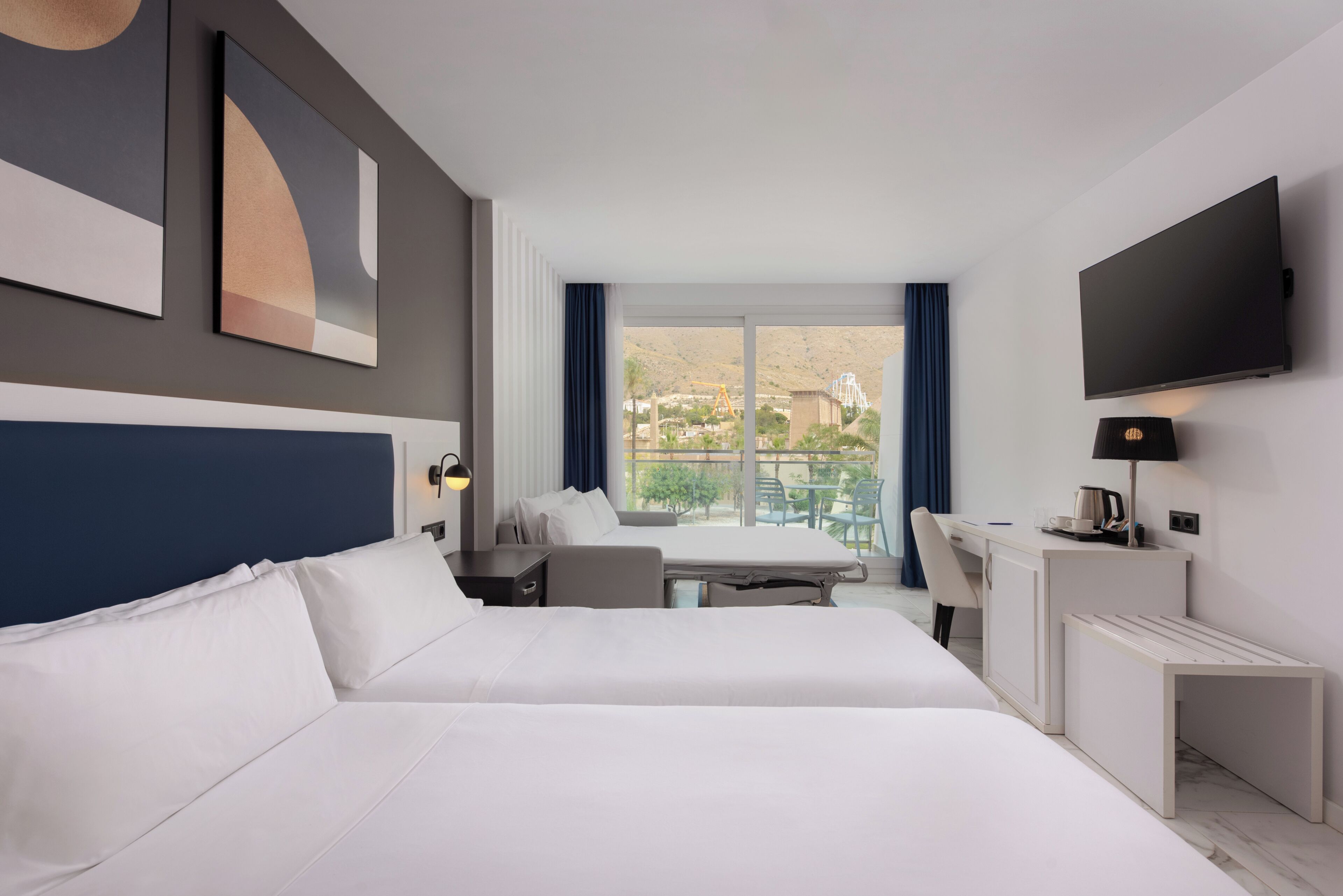 A hotel room with a bed, desk, chair, TV, and a balcony with a view.