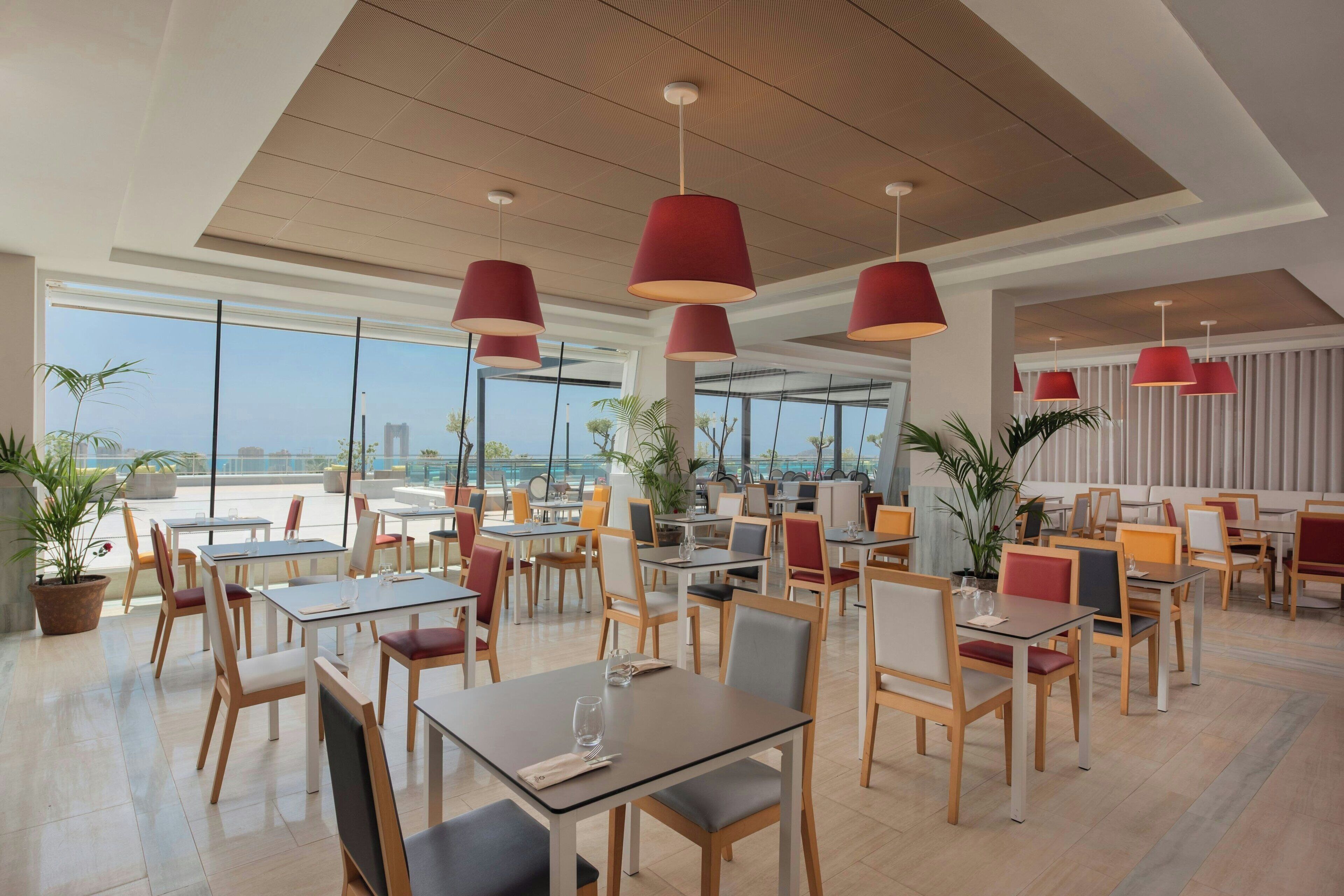 A modern dining area with tables and chairs, large windows, and red pendant lights.