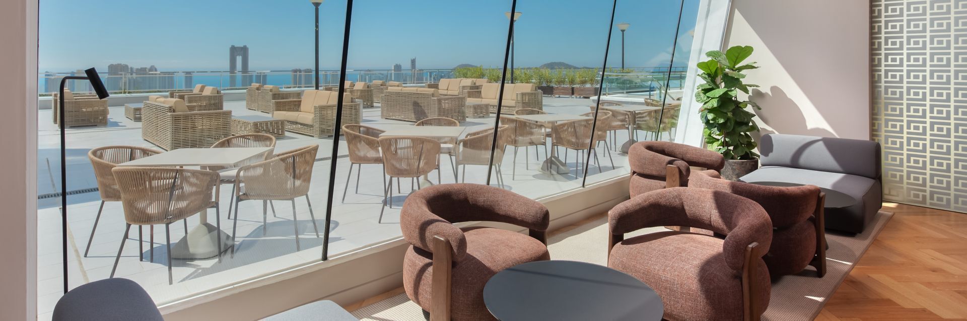 A modern lounge area with wicker furniture, a grey sofa, and a view of the city skyline through large windows.
