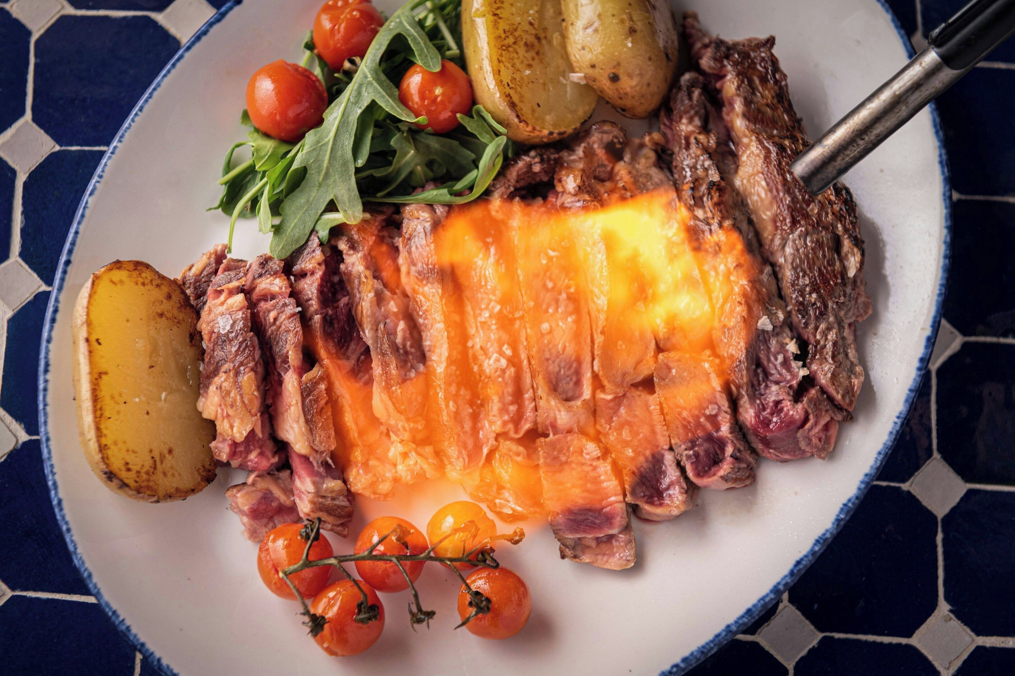 Un plato con carne en rodajas, patatas, tomates cherry y verduras de hoja verde.