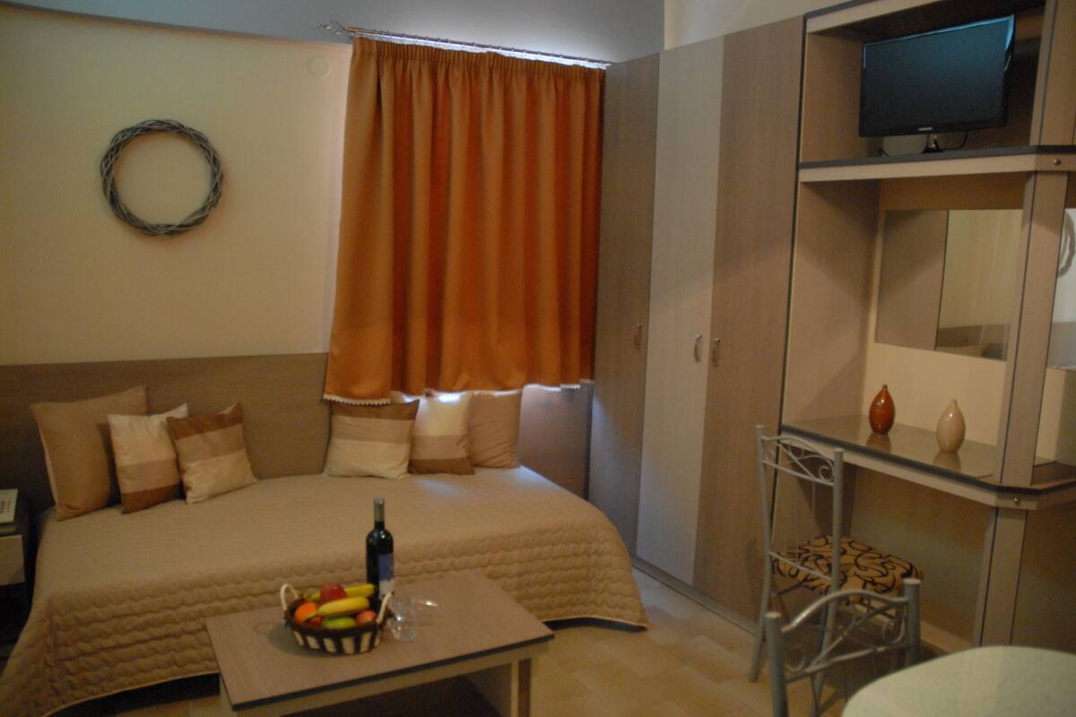A hotel room with a bed, a table with a fruit bowl, a bottle, and a glass. There is a television on a shelf above the bed.