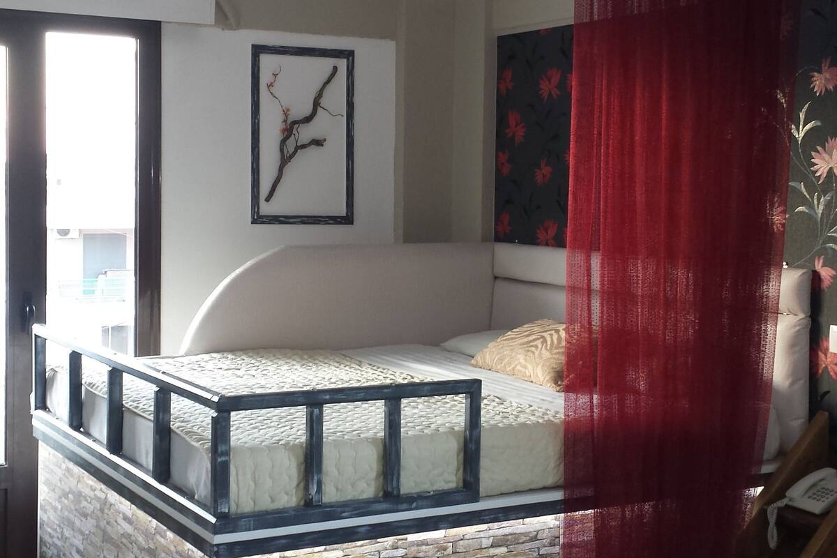 A bedroom with a glass bed platform, stone wall, a hanging light fixture, a bed with a white mattress, a red curtain, and a bedside table with a remote control.