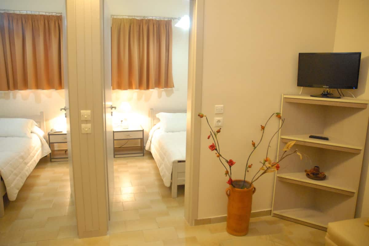A hotel room with two beds, a TV on a stand, a vase with flowers, and a shelf with a remote control.