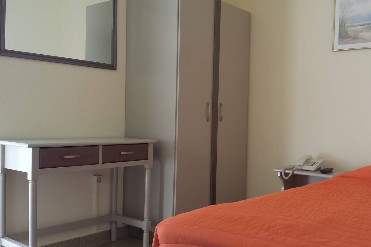 A room with a bed, a desk with drawers, a wardrobe, a telephone, and a painting on the wall.