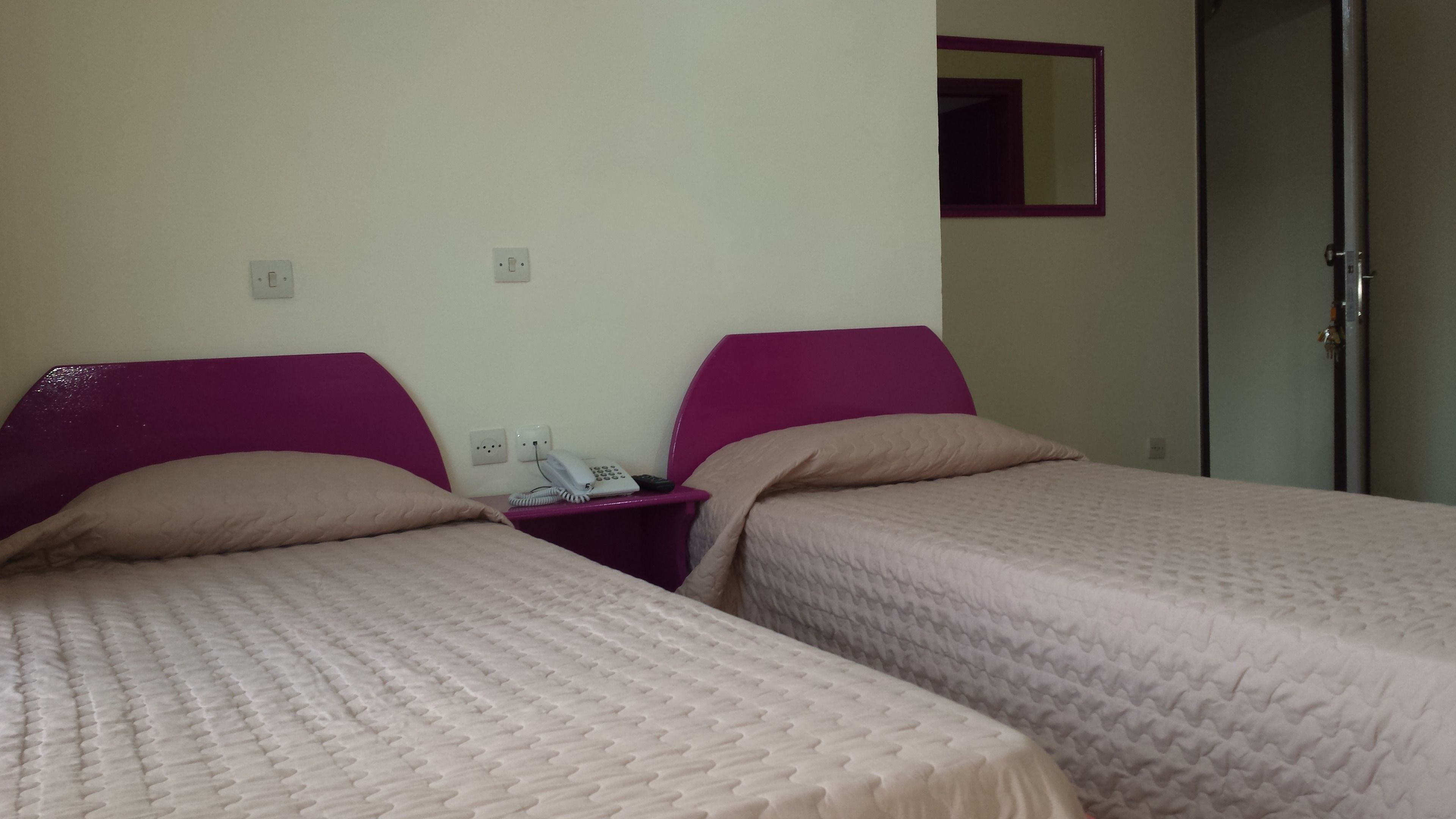 A hotel room with two beds, each with a headboard and a pillow. There is a nightstand with a phone and a remote control.