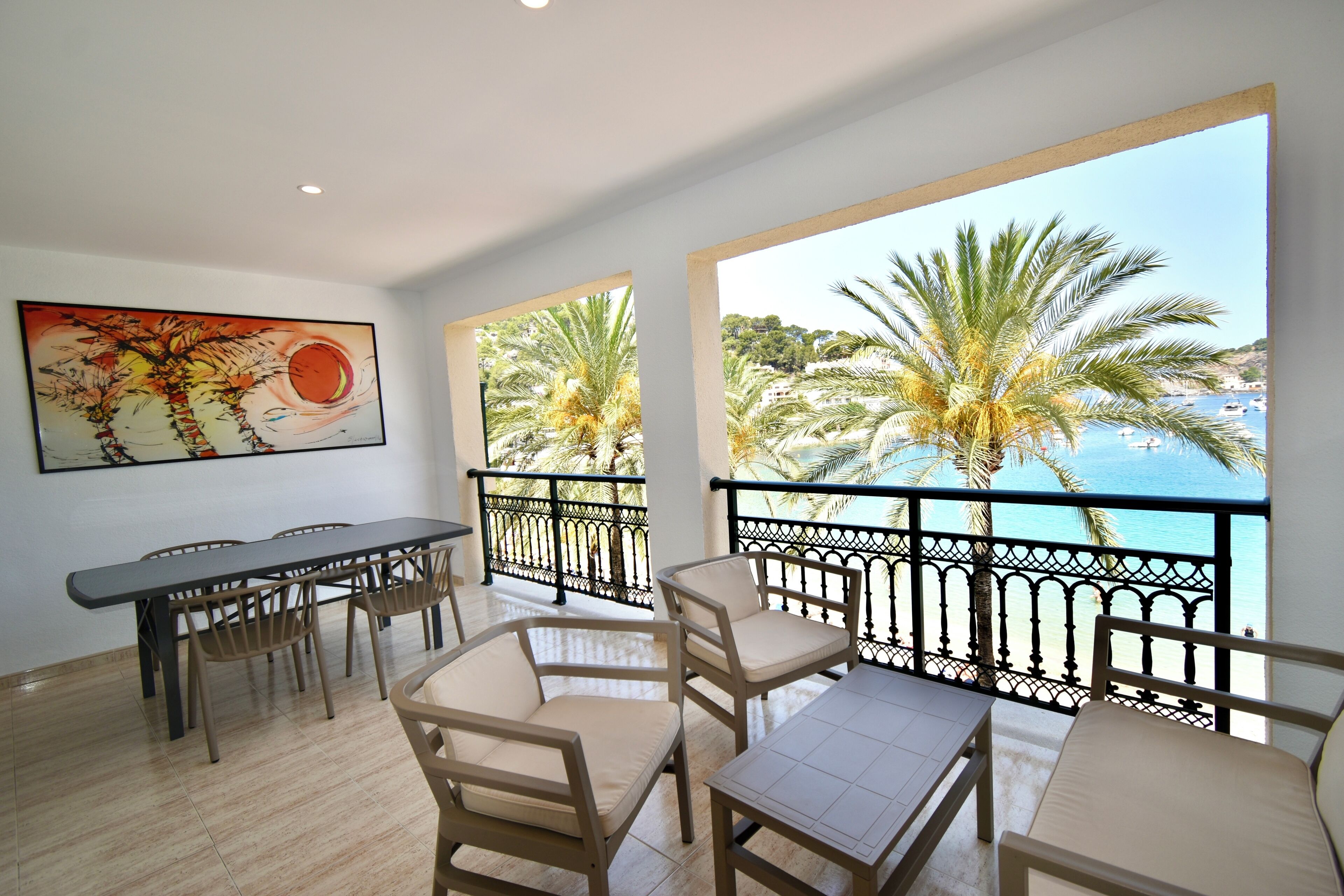 A dining area with a table and chairs, a painting on the wall, and a balcony with a view of palm trees and the sea.