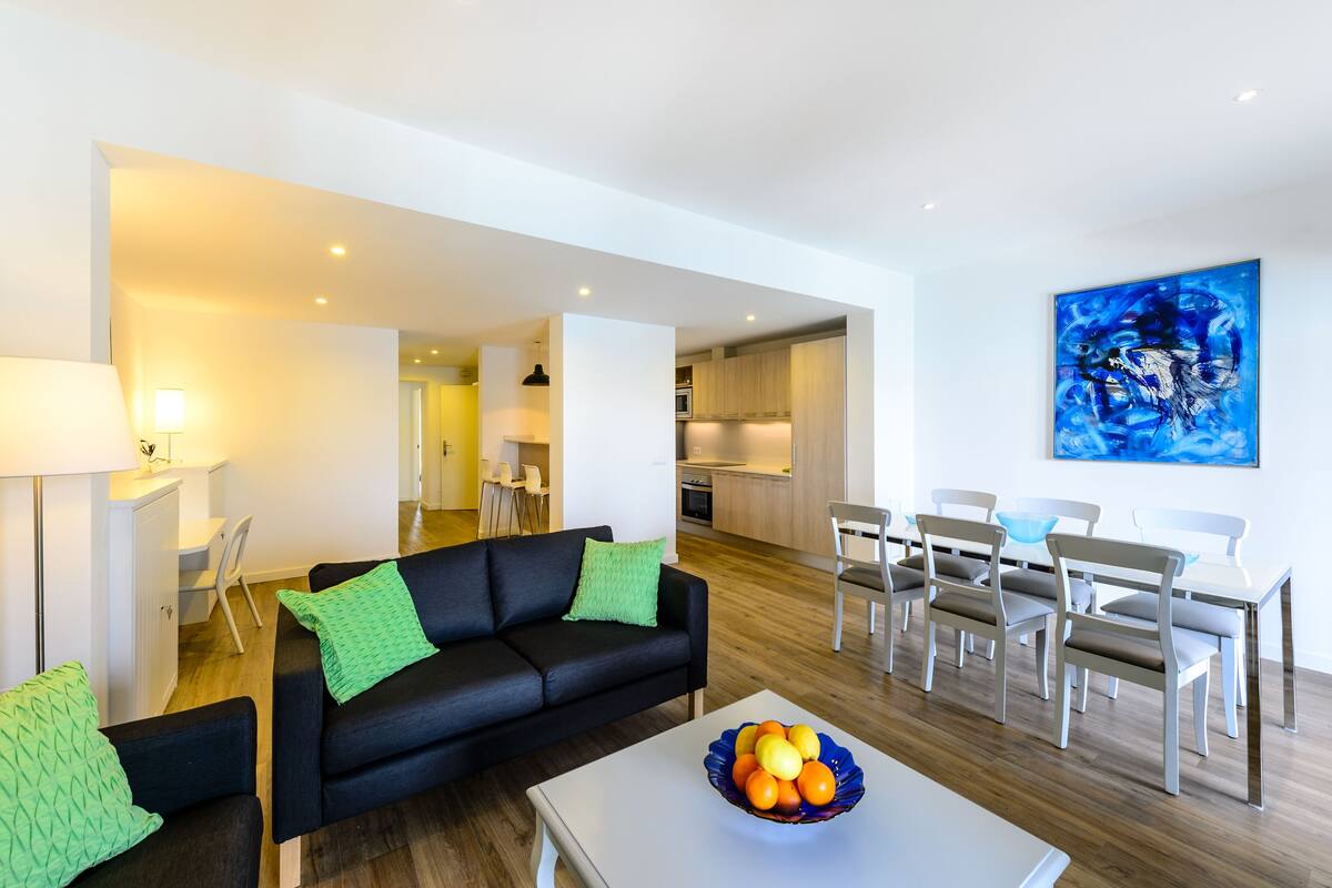 A modern living room with a black sofa, a dining table with chairs, and a kitchen in the background.