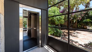 A modern elevator with a glass door, leading to a room with a view of lush greenery and a tiled floor.