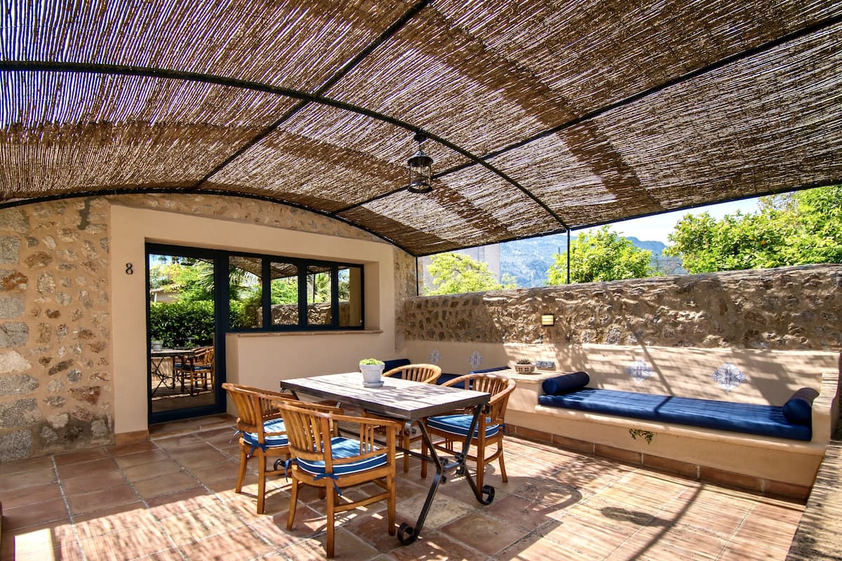 An outdoor patio with a stone wall, a wooden table with chairs, a cushioned bench, and a pergola overhead.