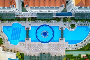 Vista aérea de un hotel con una gran piscina central, varios balcones y un área exterior bien cuidada con sillas de descanso y sombrillas.
