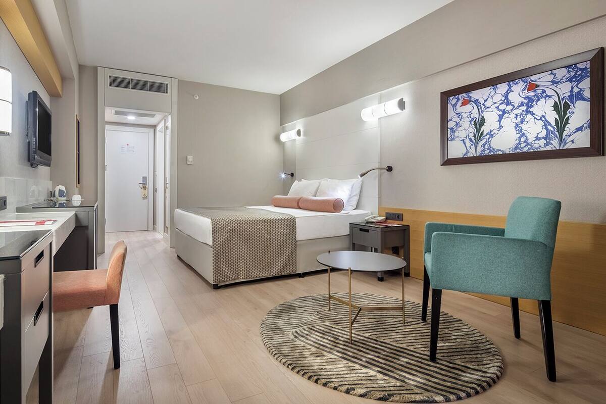 A modern hotel room with a bed, desk, chair, and a small table. There is a painting on the wall and a rug on the floor.