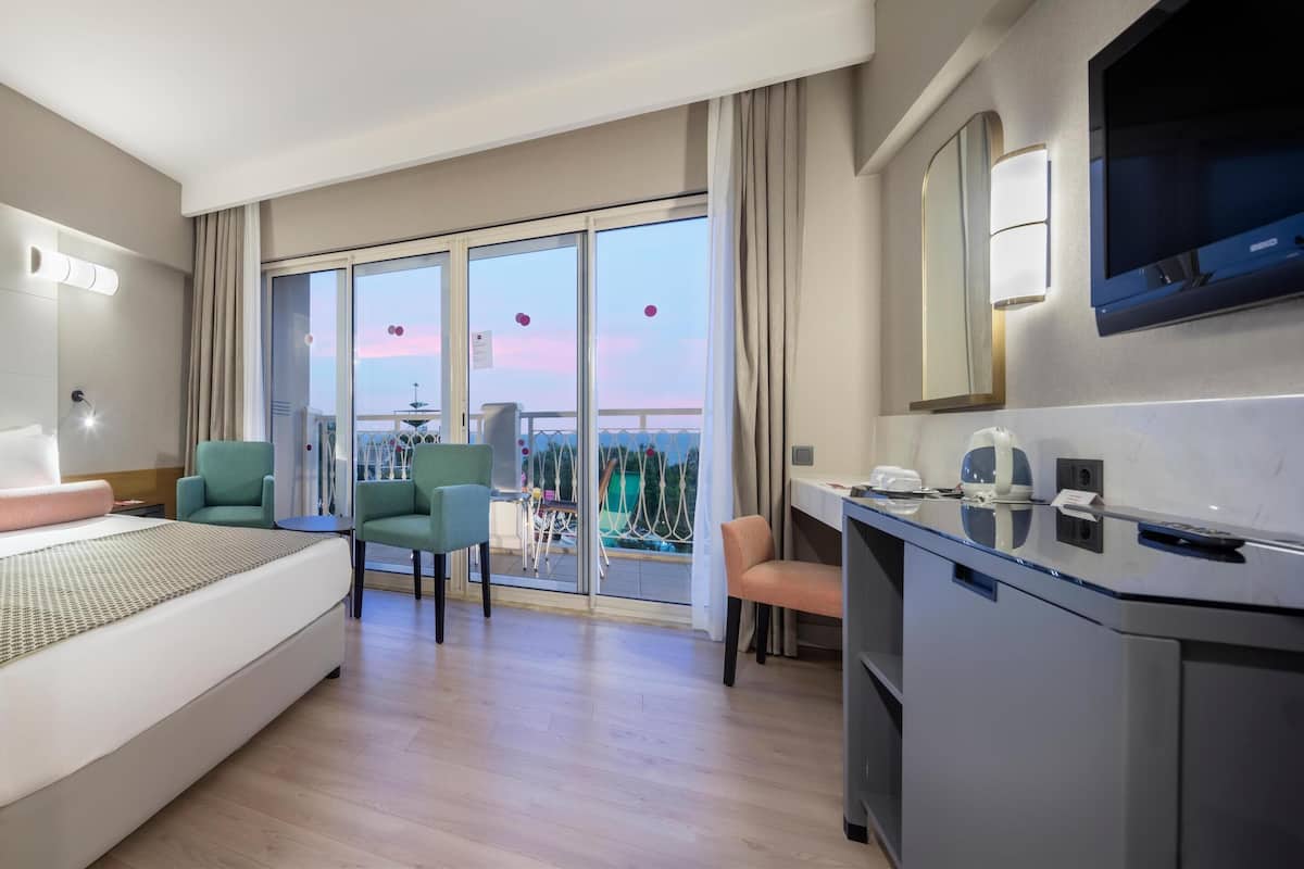 A modern hotel room with a large bed, a desk, a chair, and a balcony with a view.