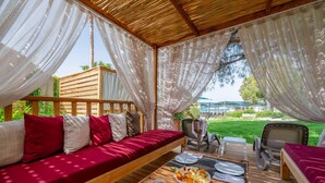 A covered outdoor seating area with a wooden bench, a table with plates of food, and a view of the garden. - Trendy Palm Beach (Adults Only, 16+) - All inclusive (Manavgat)