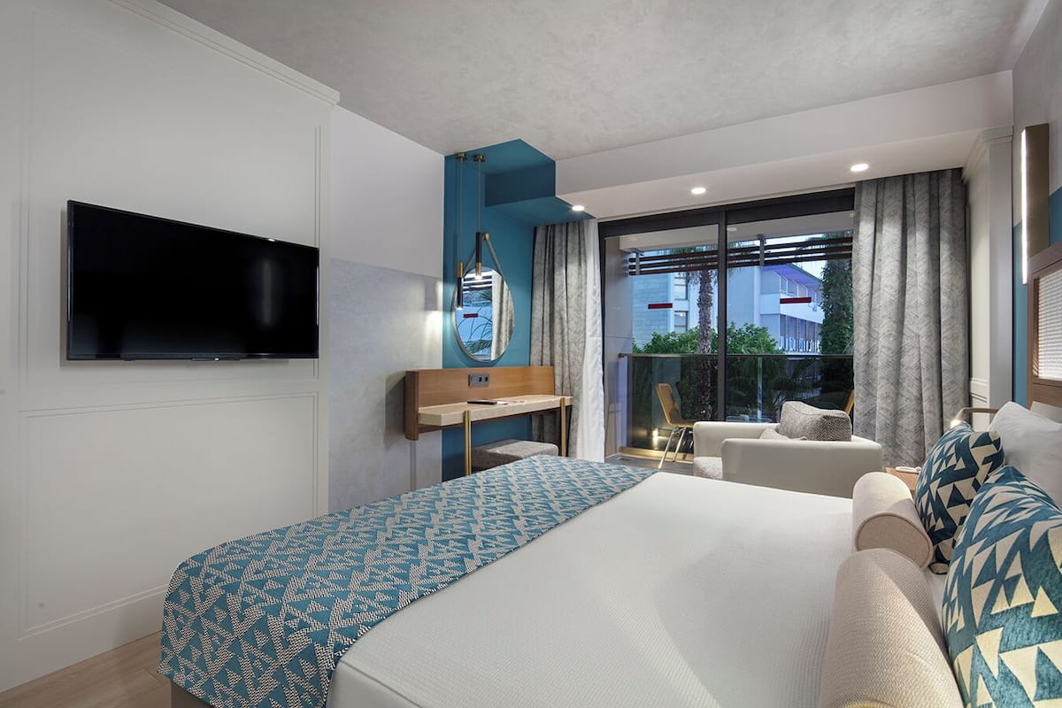 Quarto de hotel moderno com uma cama grande, televisão na parede, secretária, cadeira e vista exterior por uma janela ampla.