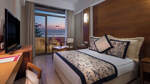 A hotel room with a bed, a desk, a TV, a phone, and a view of the sunset over the water. - Trendy Side Beach (Adults Only, 16+) - All inclusive (Manavgat)
