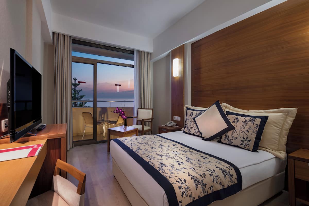 Quarto de hotel com cama, escrivaninha, televisão, telefone e vista para o pôr do sol sobre a água.