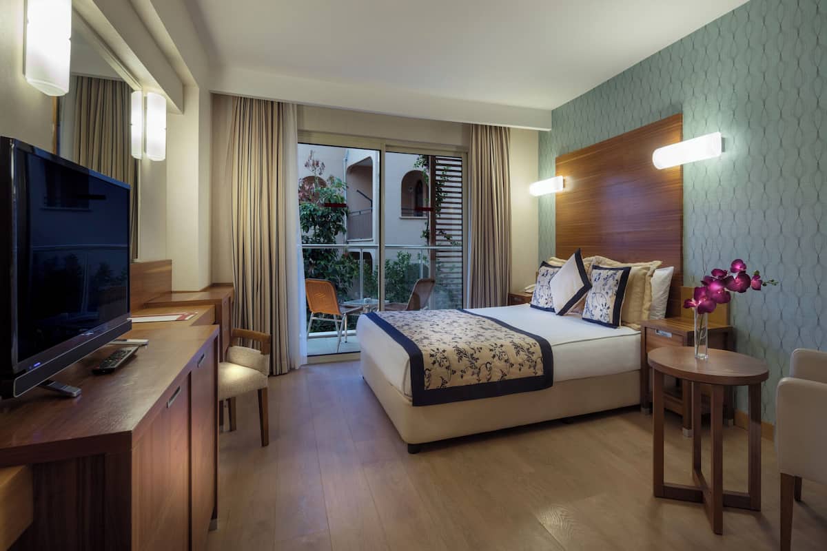 Quarto de hotel com cama, escrivaninha, televisão, cadeira e mesa pequena com um vaso de flores.