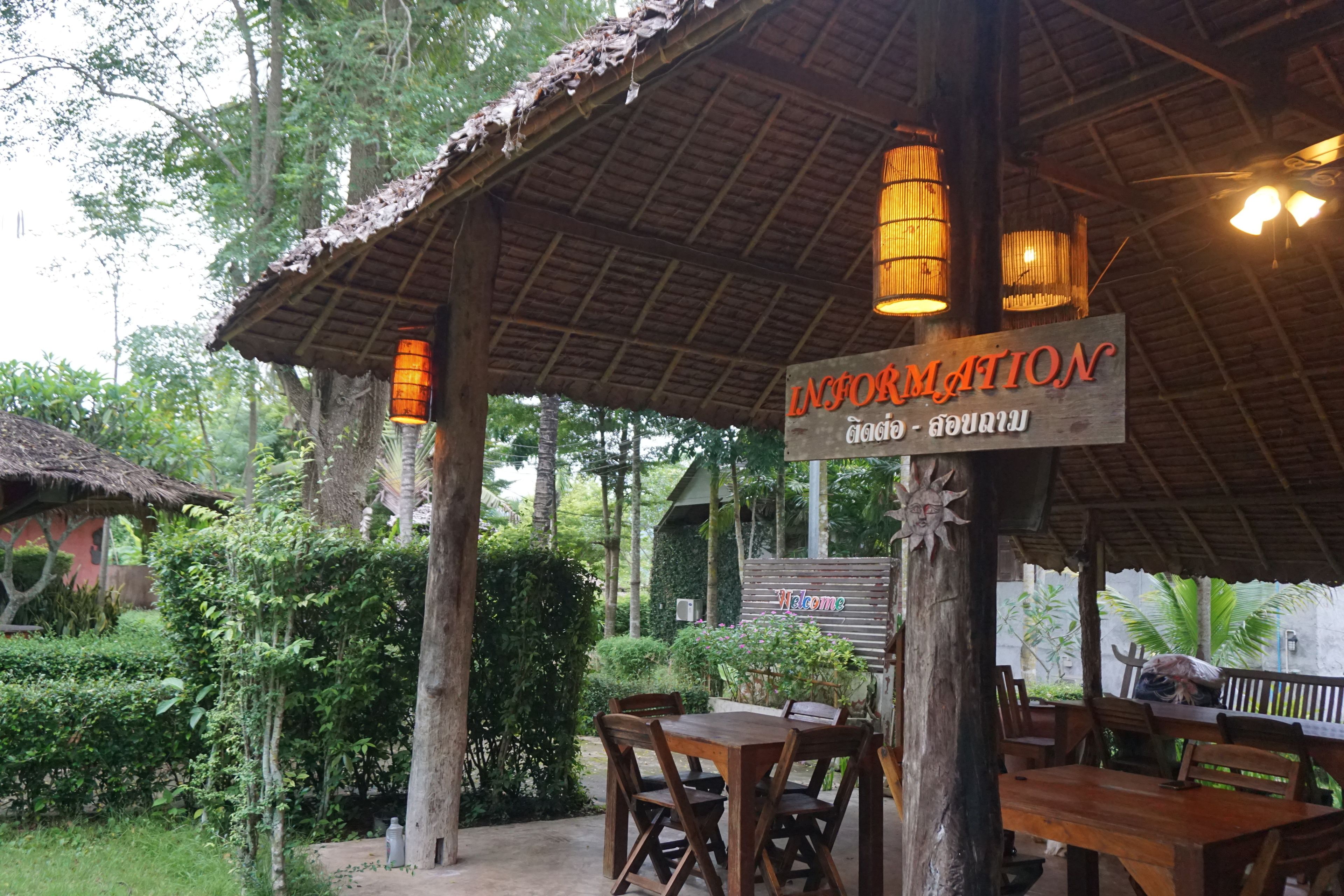 A rustic outdoor seating area with wooden tables and chairs, surrounded by lush greenery and a thatched roof structure.
