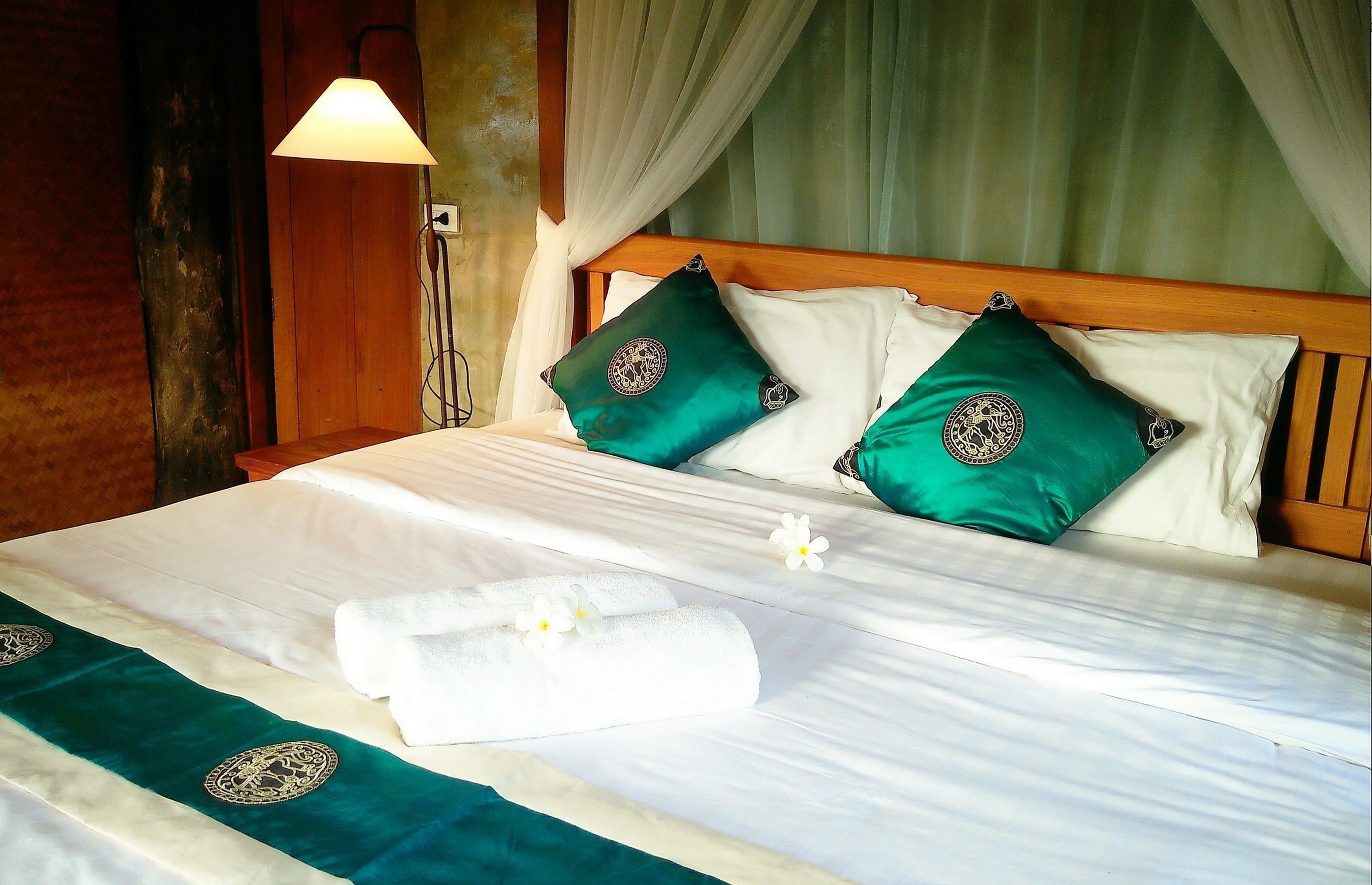 A neatly made bed with white linens, two green pillows with a circular emblem, and a white towel on the bed.