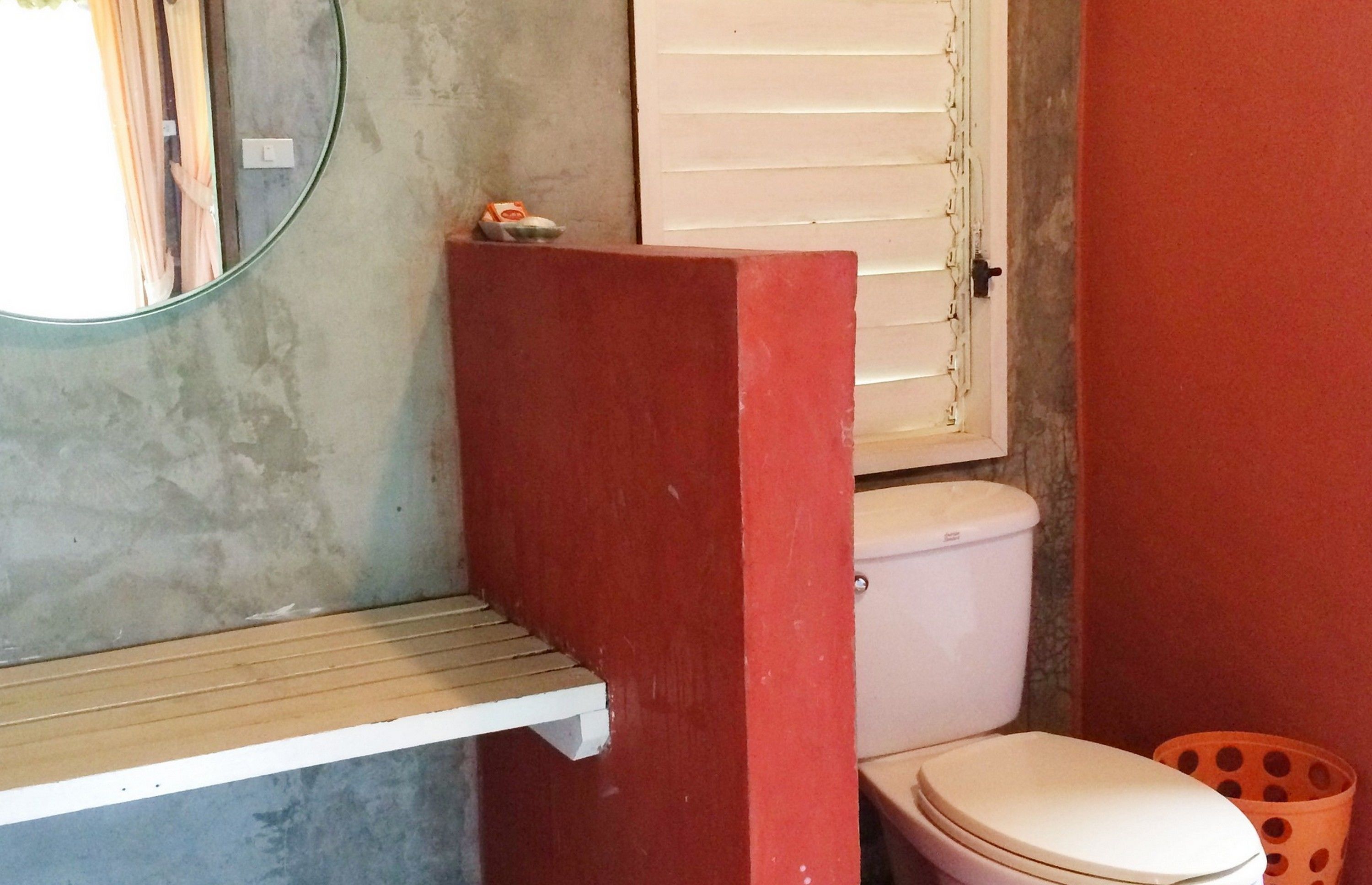 Een badkamer met een rode wand, een wit toilet, een klein plankje en een raam met zonwering.
