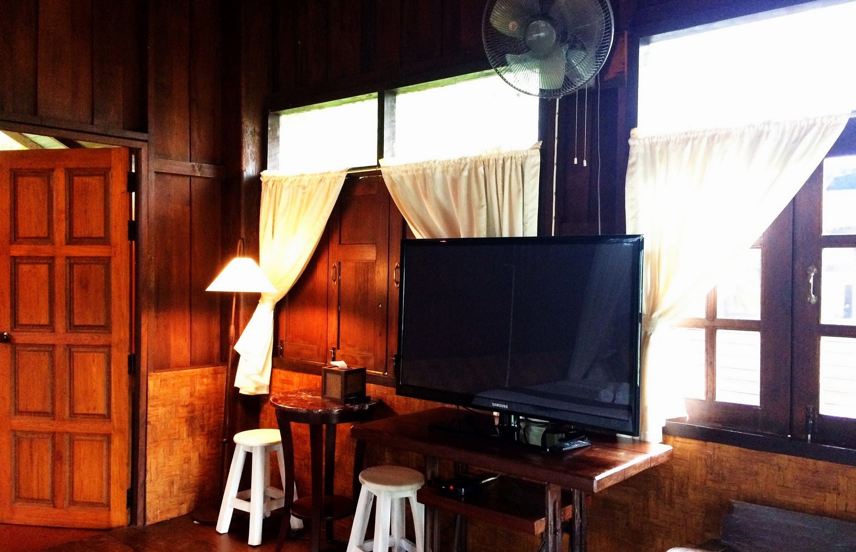 A wooden room with a TV on a wooden stand, a fan, a lamp, and a window with curtains.