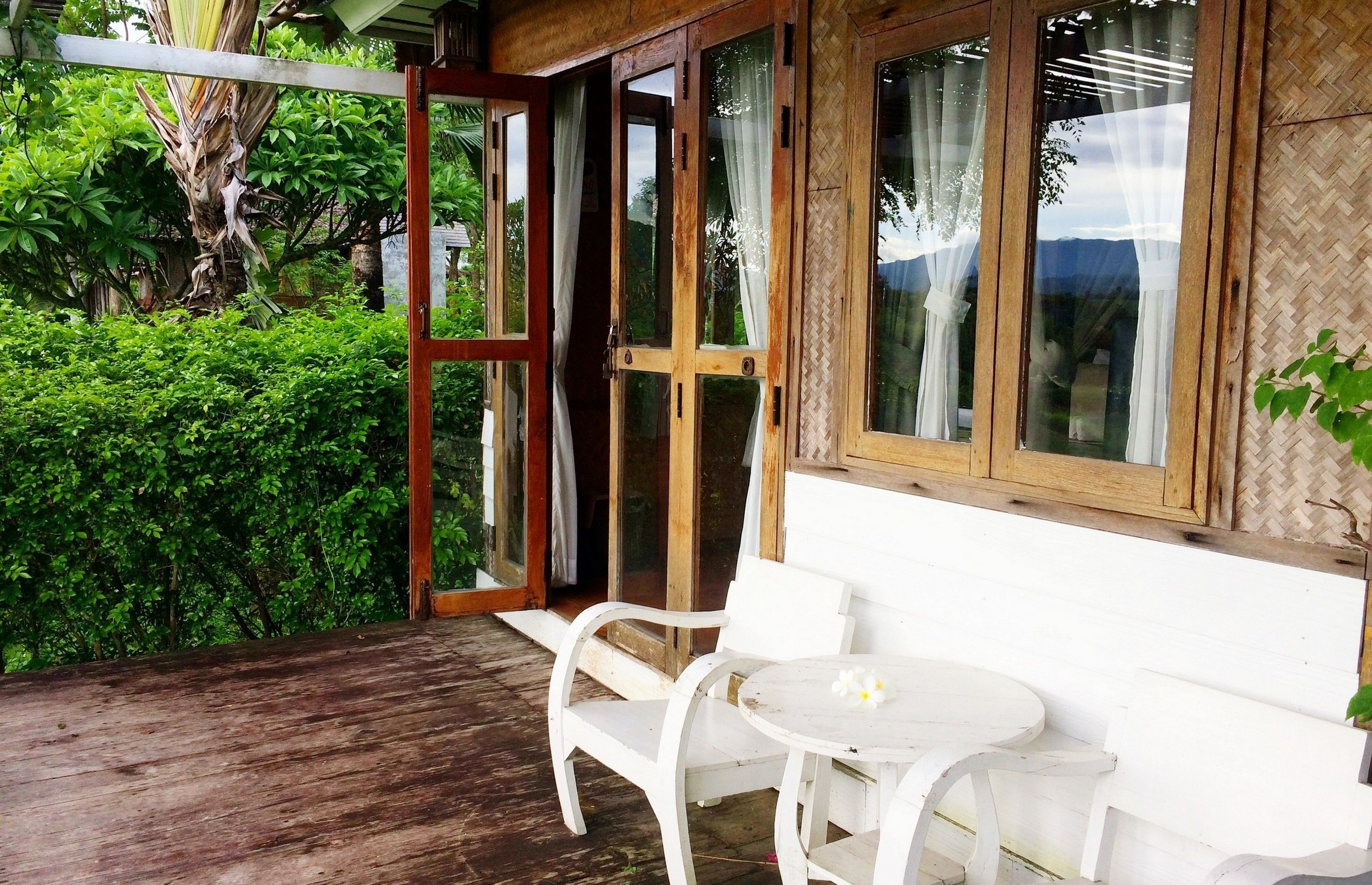 A wooden deck with white chairs and a table, a wooden door with glass panels, and a view of lush greenery.