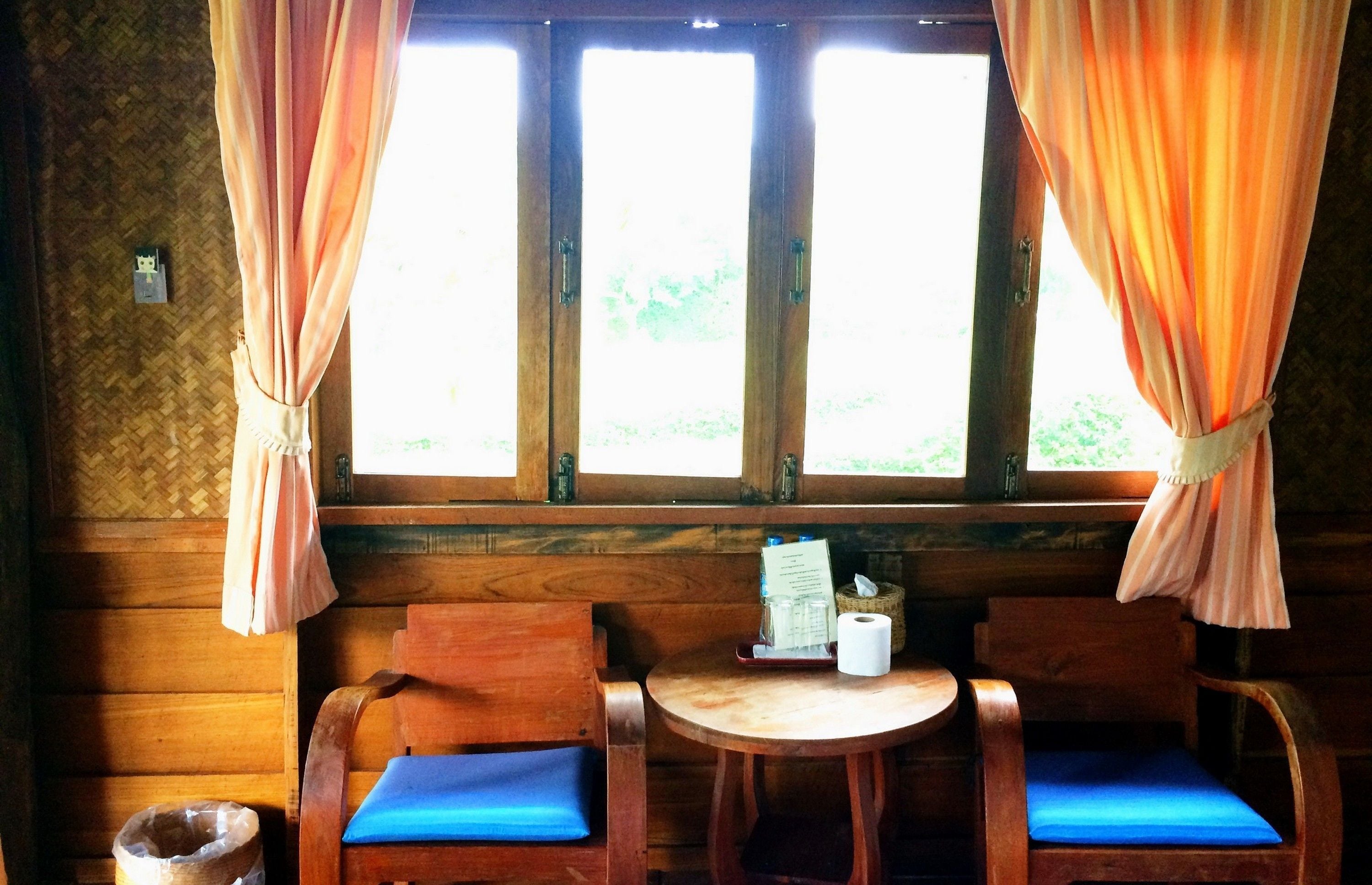 A wooden cabin with two wooden chairs with blue cushions, a small wooden table, and a window with curtains.