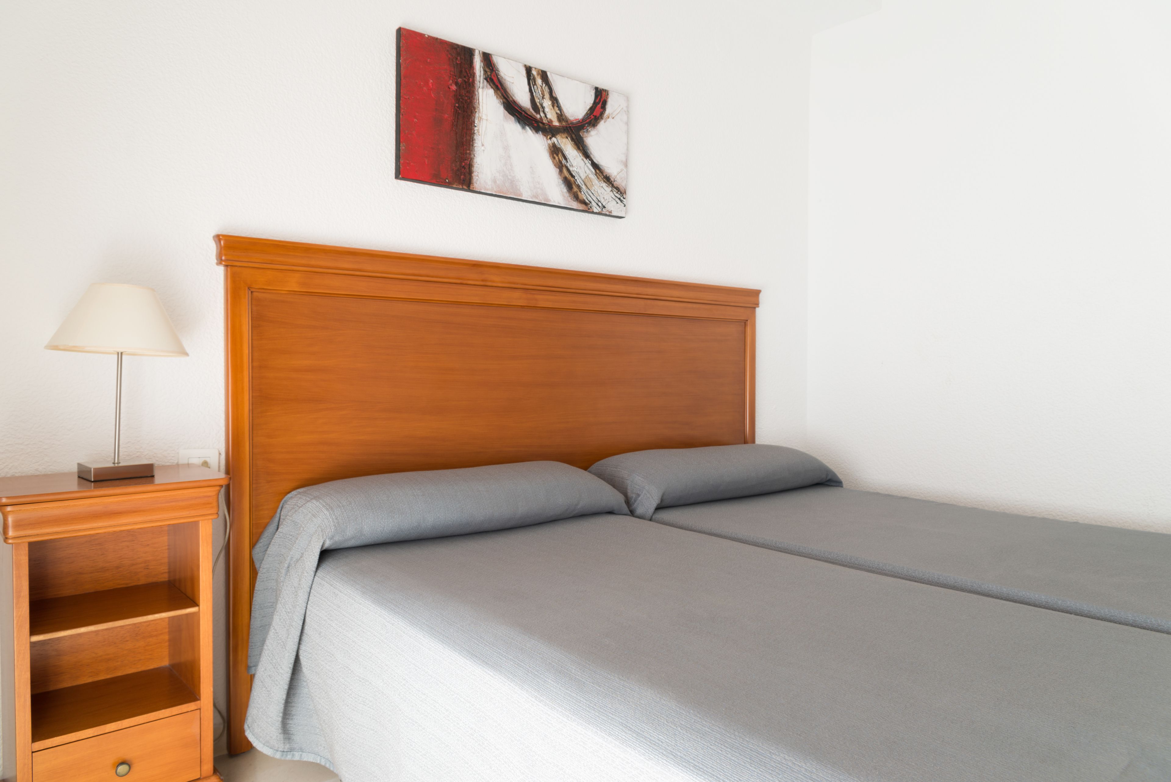 Een netjes opgemaakt bed met een houten hoofdbord, een nachtkastje met een lamp, en een abstract schilderij aan de muur.