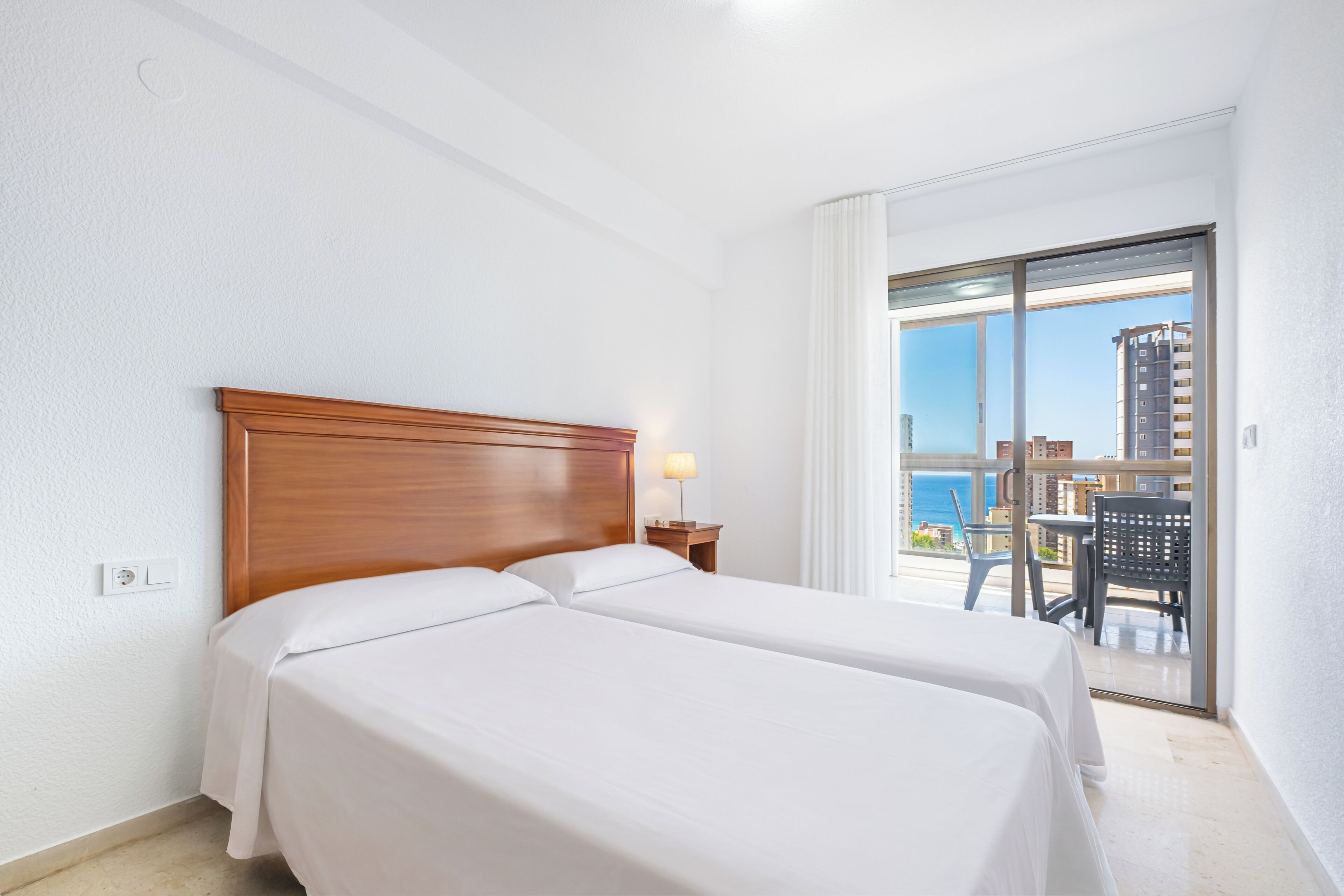 A hotel room with two beds, a nightstand, a lamp, and a balcony with a view of the sea and buildings.