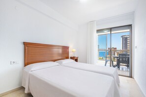 A hotel room with two beds, a nightstand, a lamp, and a balcony with a view of the sea and buildings. - Apartamentos Gemelos 2 - Beninter (Benidorm)
