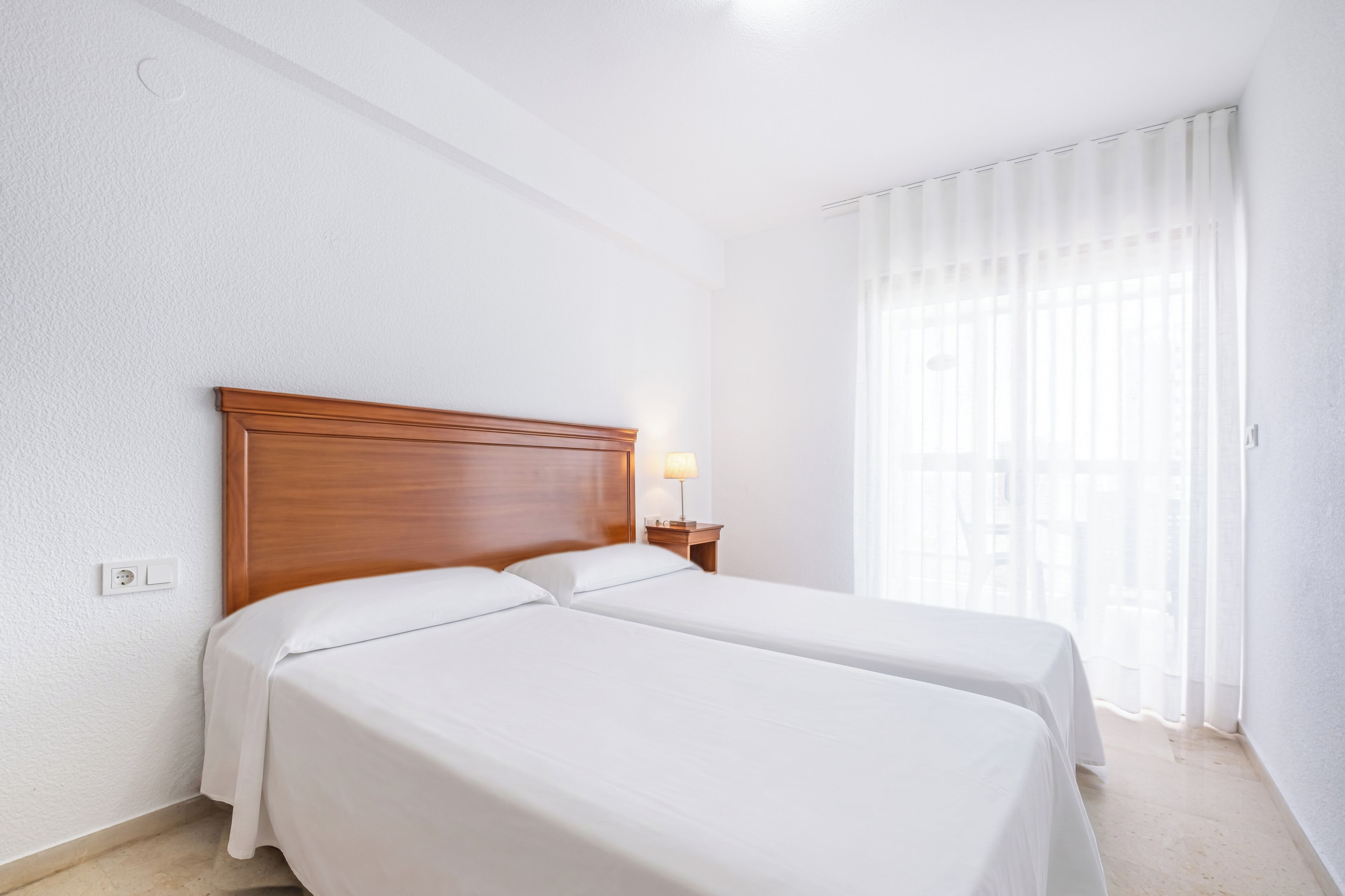 Habitación de hotel con dos camas individuales, cabecera de madera, mesita de noche con lámpara y ventana con cortinas translúcidas.