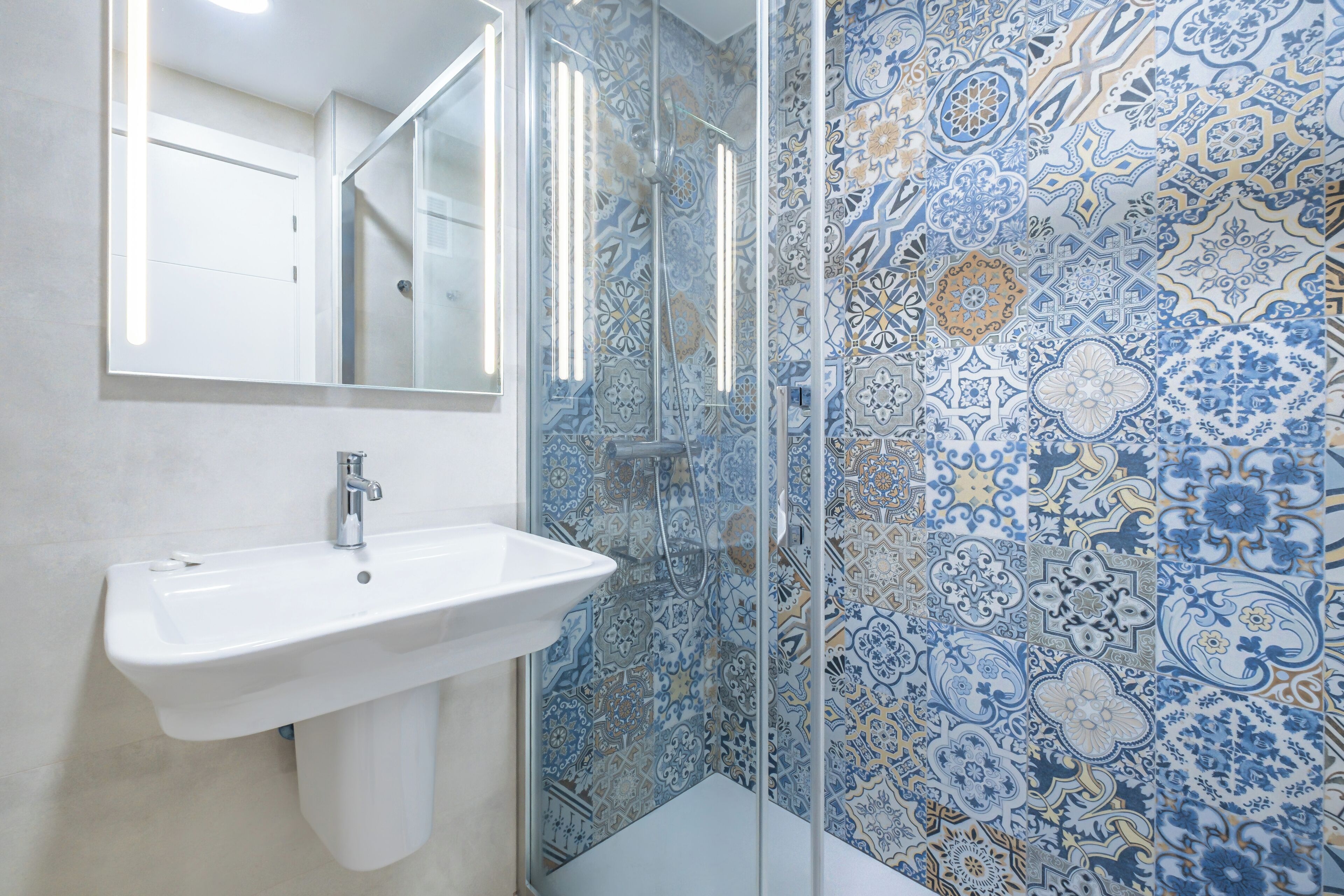 Un bagno con un lavandino bianco, cabina doccia in vetro e parete rivestita di piastrelle con motivi elaborati.