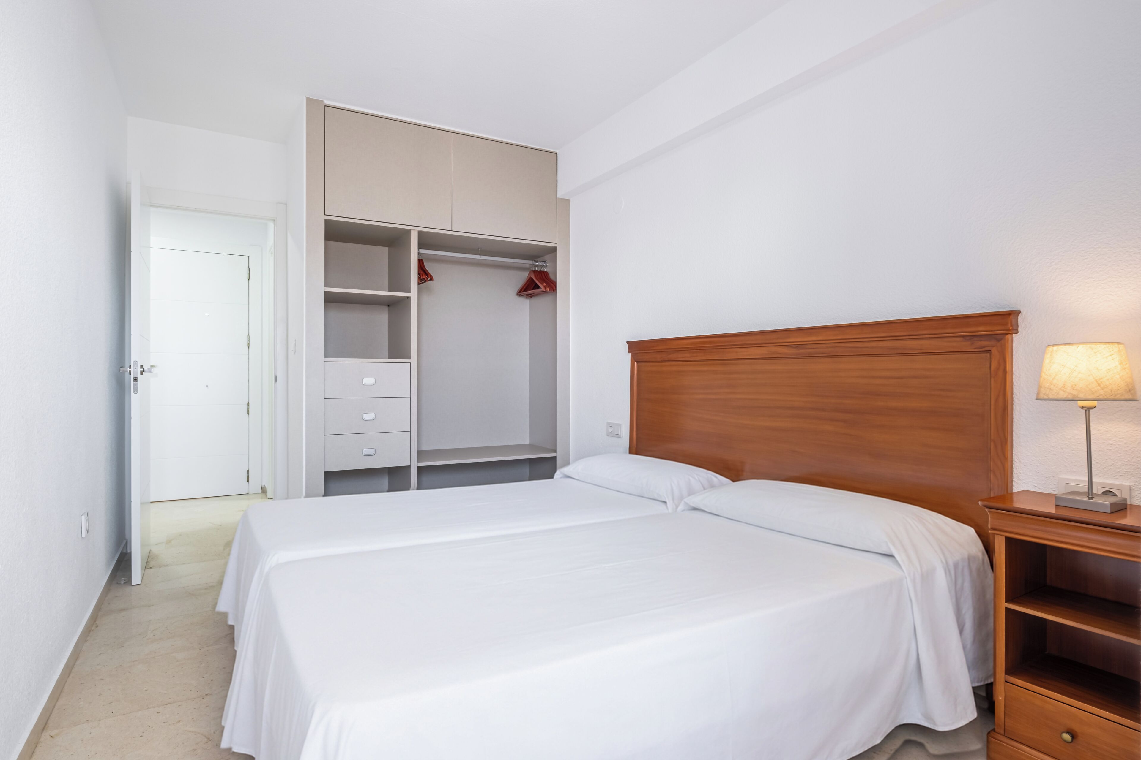 Een slaapkamer met een tweepersoonsbed, een houten hoofdbord, een nachtkastje met lamp, een kledingkast met hangruimte en een deur die naar een andere kamer leidt.