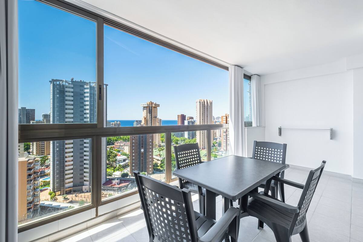 Un balcón con mesa y sillas, ventanas amplias con vista a la ciudad y un interior en tonos blancos.
