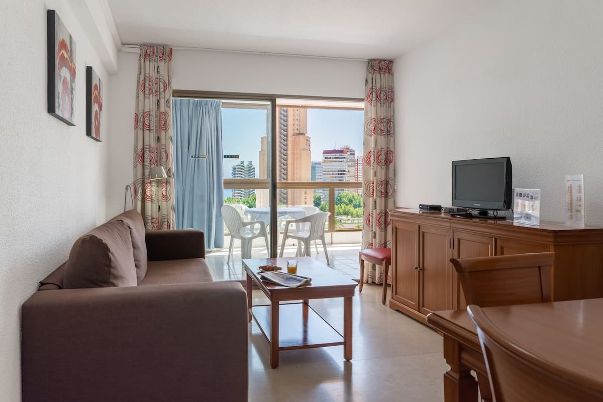 Una sala de estar con sofá, una mesa de centro, televisor y un balcón con vistas a edificios.