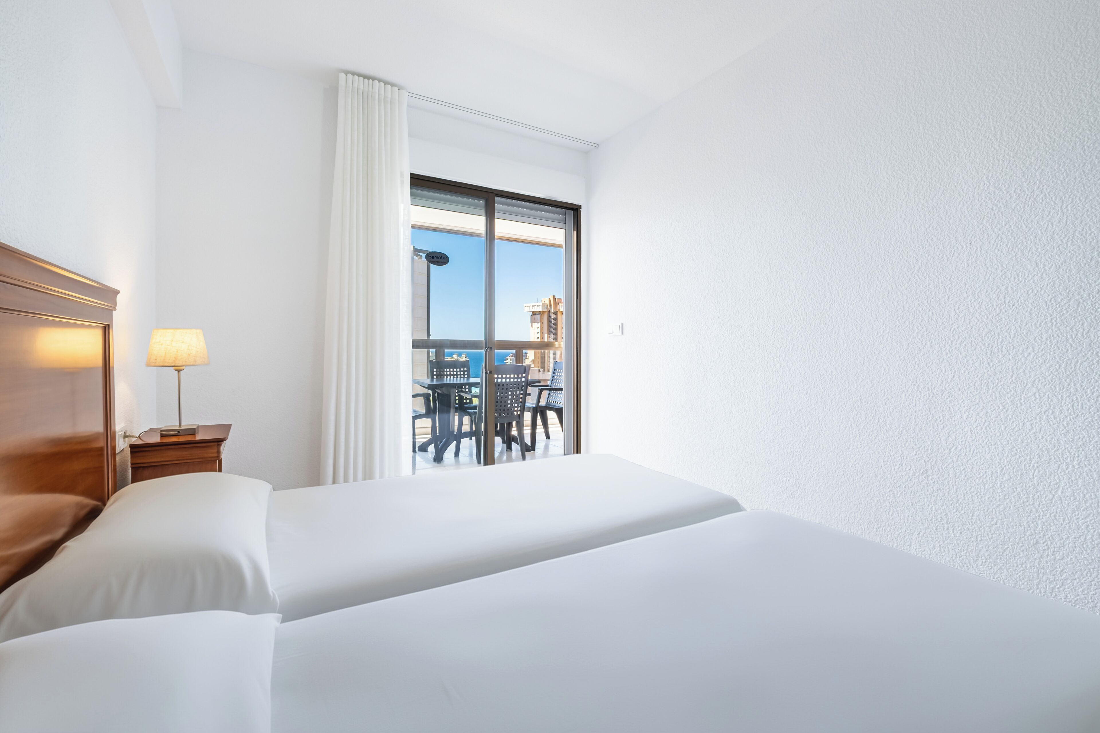 A hotel room with two beds, a nightstand with a lamp, a balcony with a table and chairs, and a view of the cityscape.