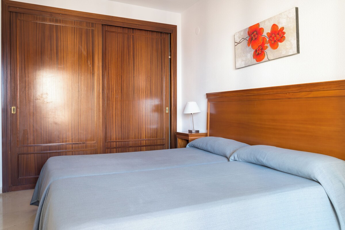 A bedroom with a wooden wardrobe, a bed with grey bedding, and a wall-mounted artwork of flowers.