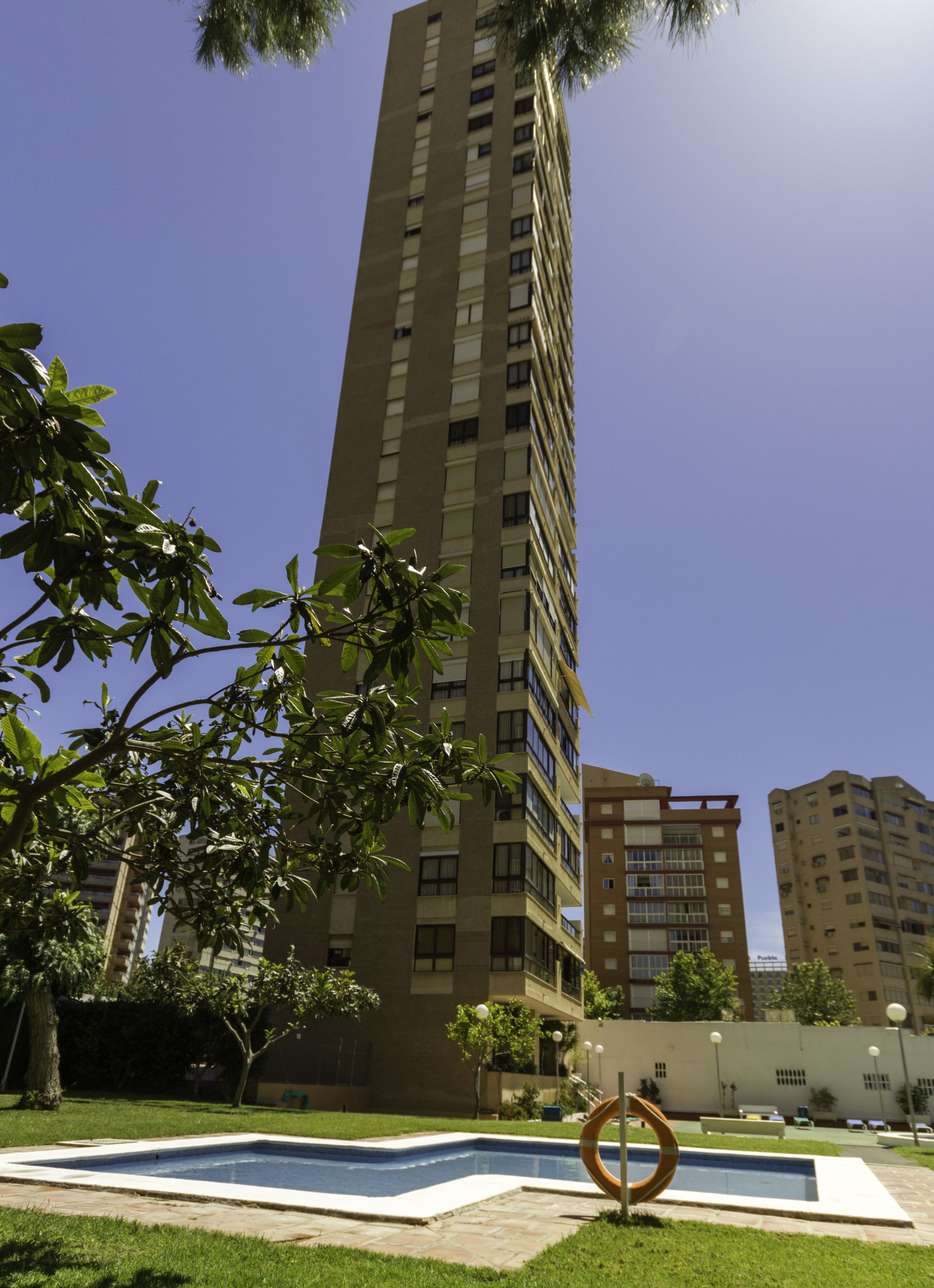 A tall residential building with multiple balconies and windows, surrounded by greenery and a swimming pool area.