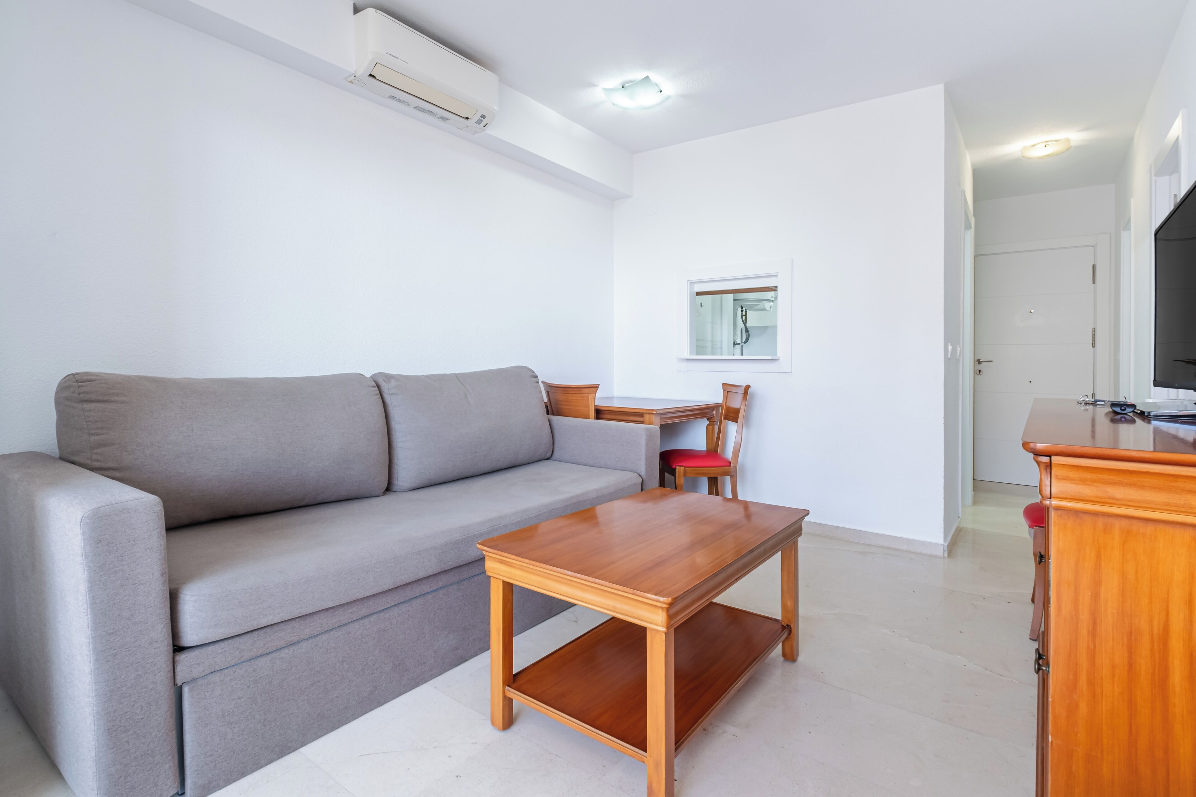 Un salón con un sofá gris, una mesa de centro de madera, un mueble para televisor y una ventana con persianas.