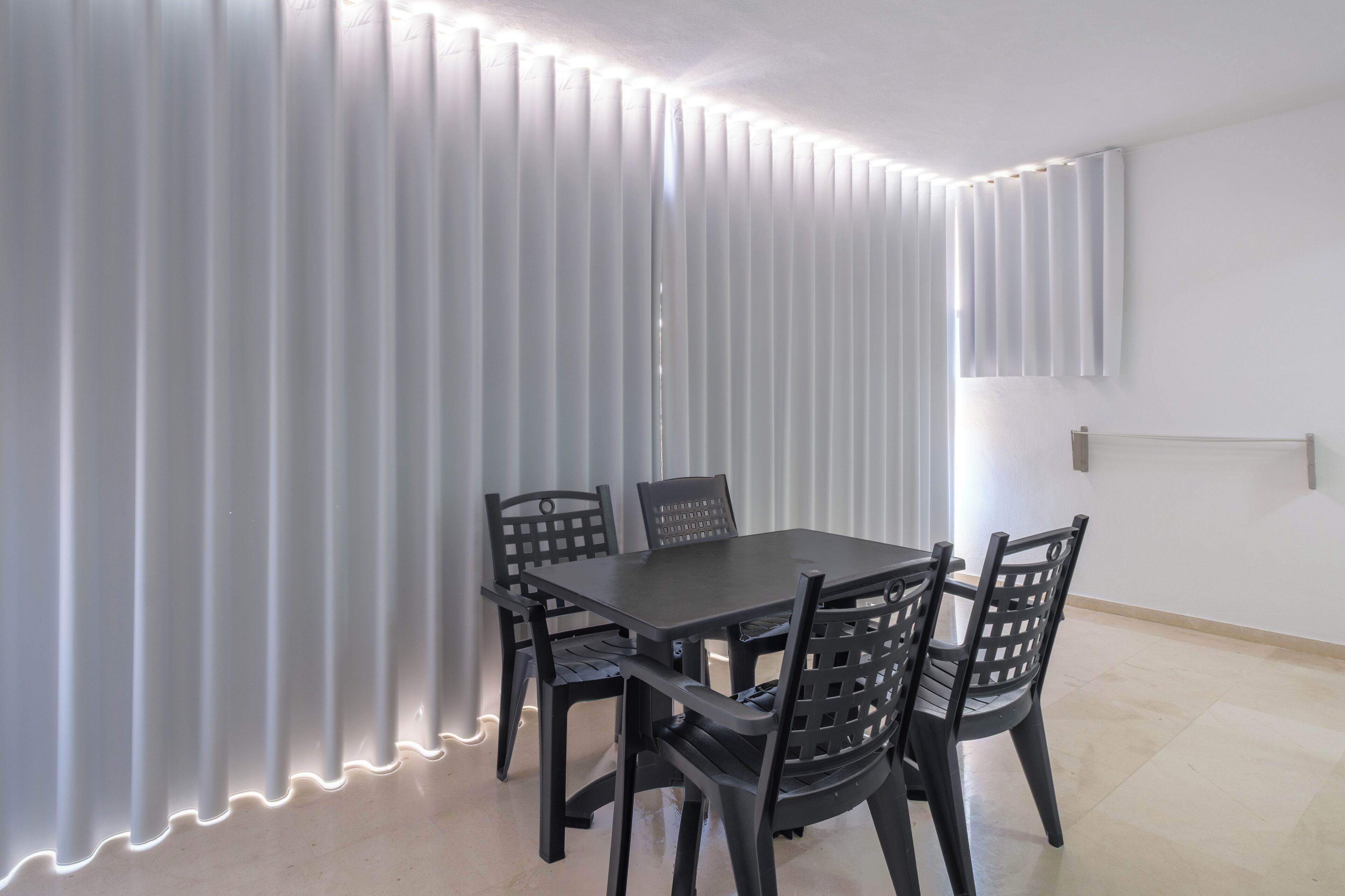 Un coin repas avec une table et des chaises, un mur blanc et une fenêtre avec des stores.