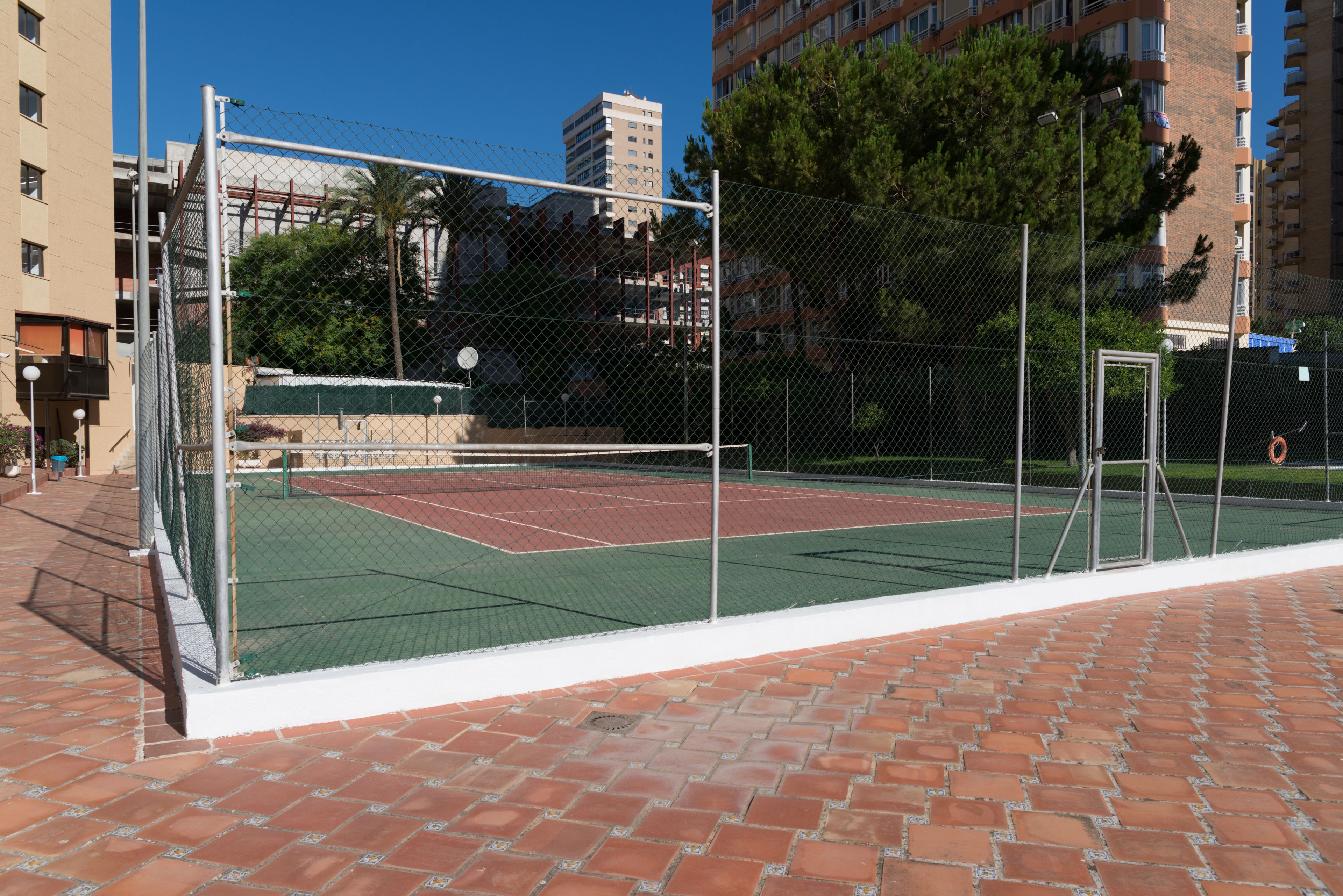 An outdoor tennis court with a chain-link fence, surrounded by a brick walkway.