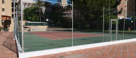 An outdoor tennis court with a chain-link fence, surrounded by a brick walkway.