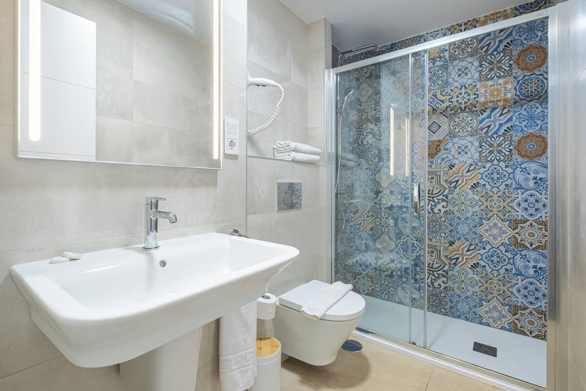 Ein Badezimmer mit weißem Waschbecken, Toilette und einer Duschkabine aus Glas mit gemusterten Fliesen.
