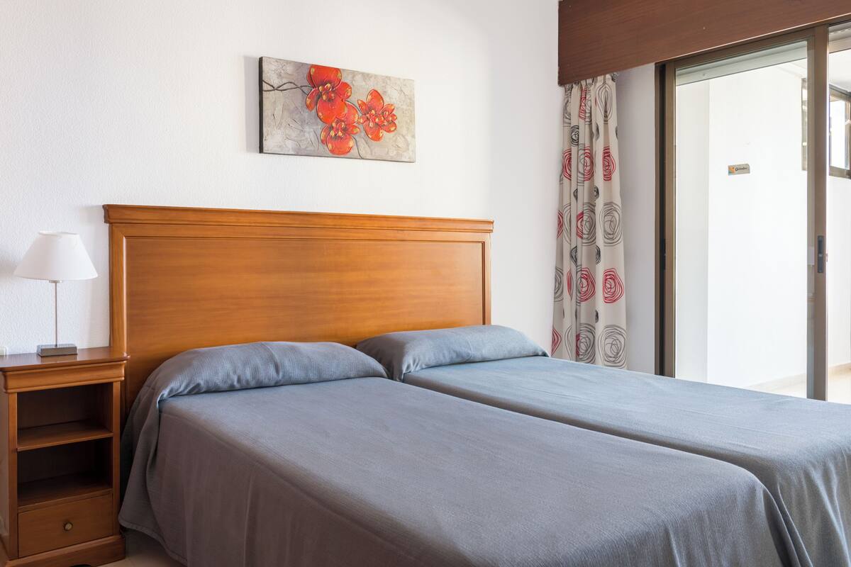 Ein Doppelbett mit grauer Bettwäsche, ein Holz-Kopfteil, ein Nachttisch mit Lampe und ein Blumenbild an der Wand.