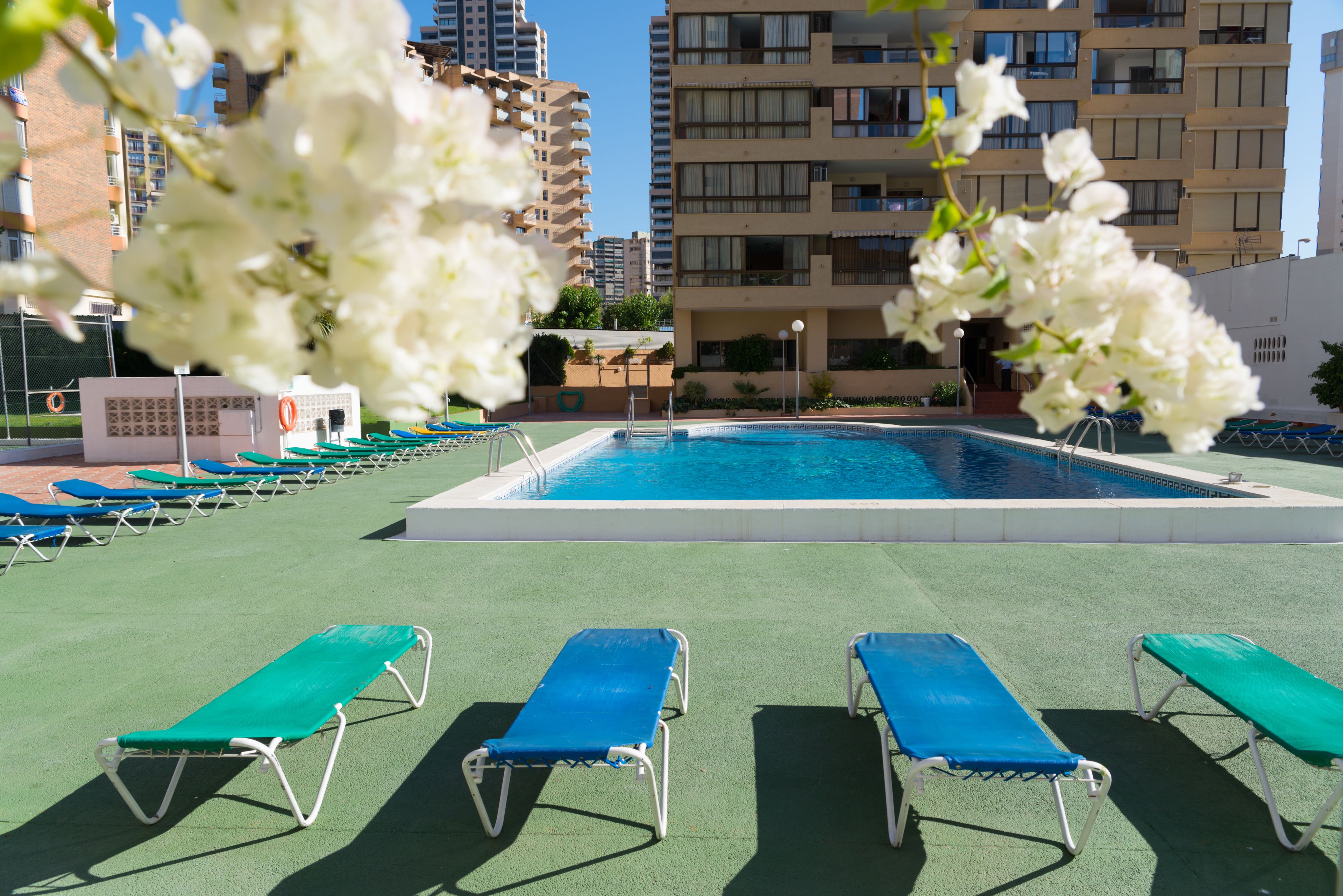 A swimming pool with blue and green lounge chairs, surrounded by a fence and a building in the background.