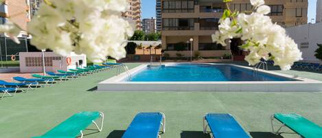 A swimming pool with blue and green lounge chairs, surrounded by a fence and a building in the background.