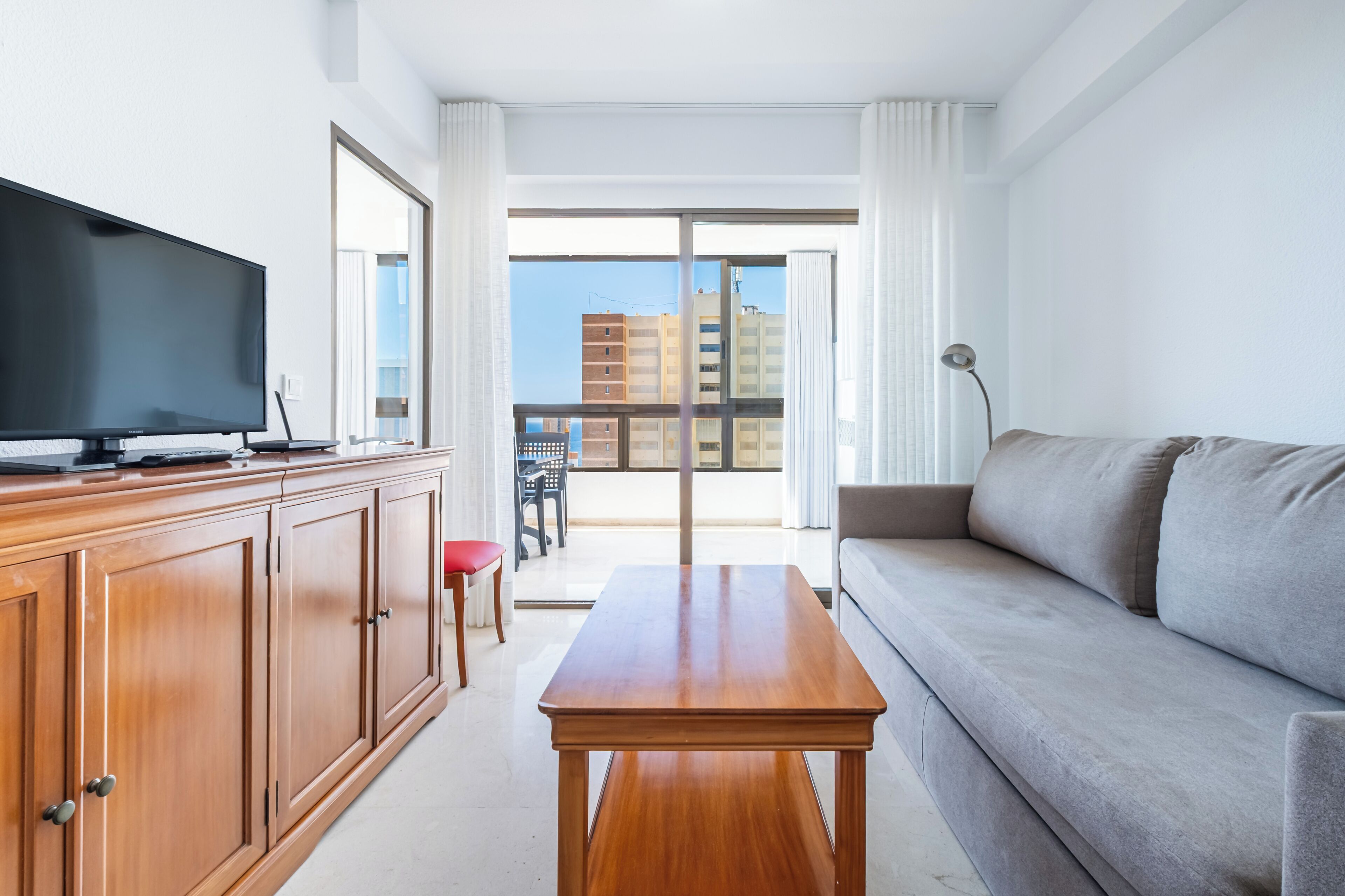 Une chambre d’hôtel moderne dotée d’une télévision à écran plat, d’une armoire en bois, d’un canapé gris et d’un balcon offrant une vue sur la ville.